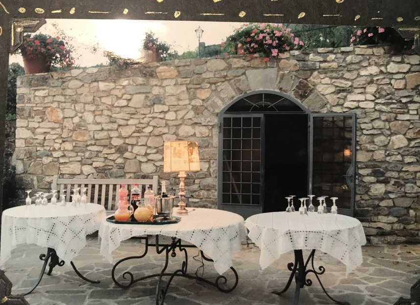 Three small round tables with white cloths, glasses, and a lamp, set against a stone wall with an arched door.