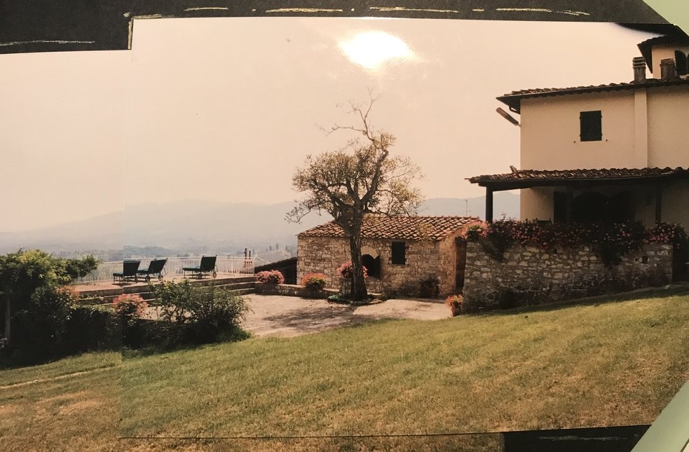 A stone outbuilding with a terracotta roof, a lone tree, chaise longues, and hills in the distance.