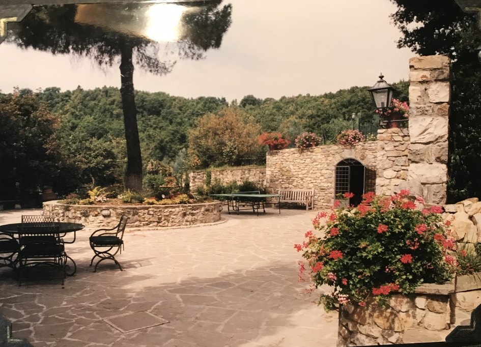 A stone courtyard with geraniums, a ping-pong table, and woods beyond a low stone wall.
