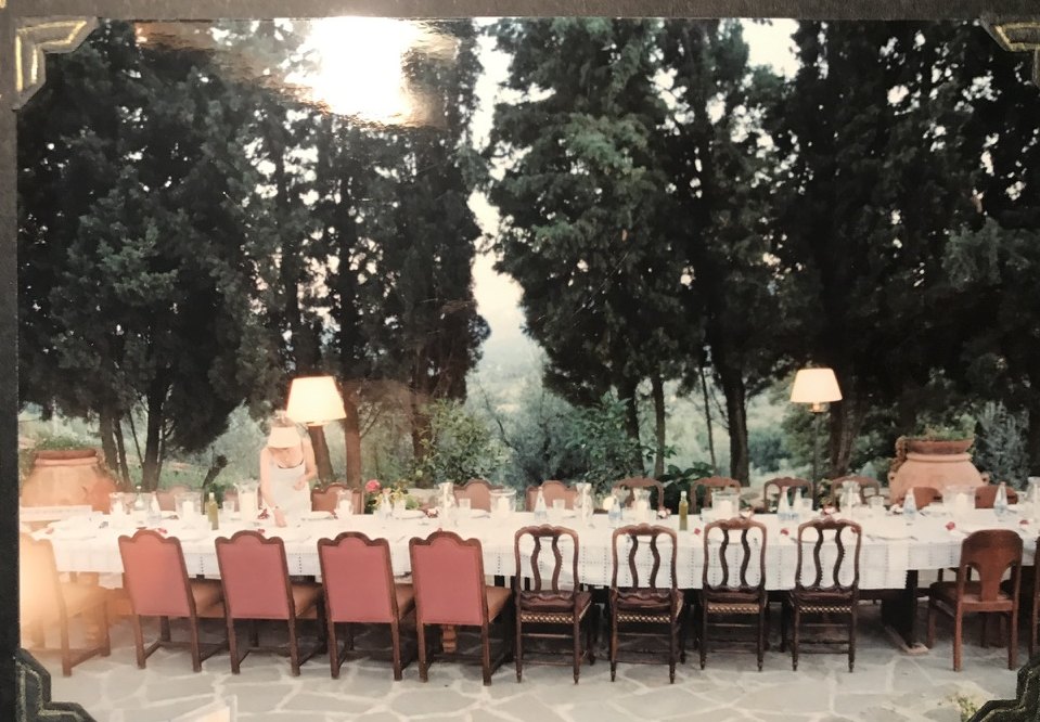 A long dining table set with a white cloth and many chairs under tall dark trees, with the valley beyond.