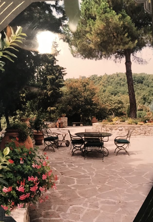 A stone patio with a round table and chairs, terracotta pots, and a tall pine.
