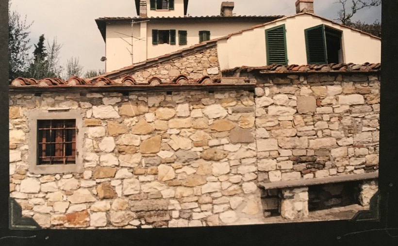 A pale stone wall with a small barred window, and the colombaia tower rising behind the roofs.