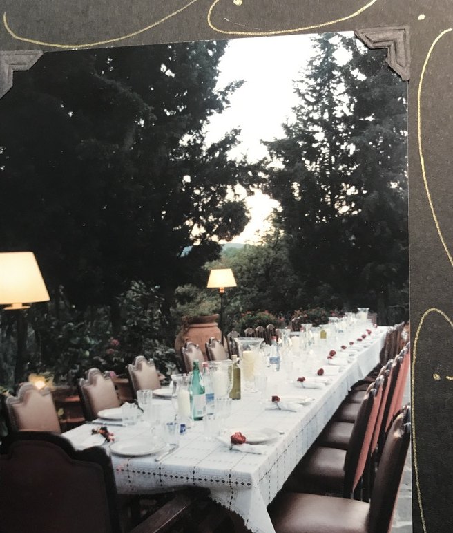 The long outdoor table seen from one end in the evening, set with glasses and bottles.